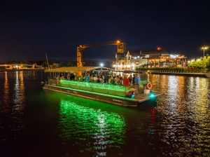 Night scene of a lit-up party barge on a river near a lit waterfront.