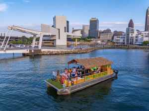 A tiki-themed boat with people onboard cruises near an urban waterfront on a clear day.