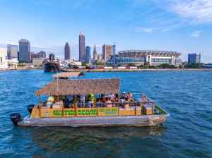 Tiki barge on water with people, city skyline and stadium in background on a clear day.