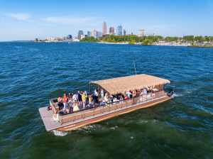 Tiki boat on lake with people, city skyline in background, sunny day.