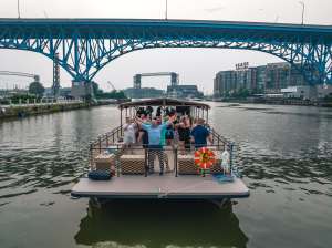 People on a party boat under a blue bridge in an urban river setting.