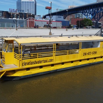 Yellow water taxi docked at a riverbank with cityscape and bridge in the background.