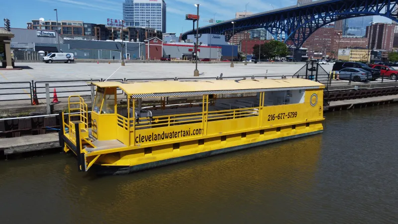 Yellow water taxi docked at a riverbank with cityscape and bridge in the background.