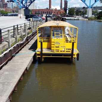 Yellow boat docked on a river next to a concrete pier with buildings and a bridge in the background.