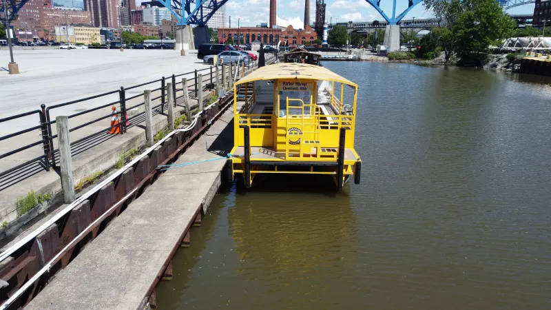 Yellow boat docked on a river next to a concrete pier with buildings and a bridge in the background.