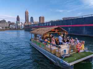 Party boat with people on a river near Cleveland skyline and docked ship.