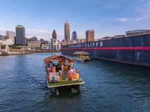 City skyline with two tiki boats on the water, large ship labeled Cleveland Cliffs in the background.